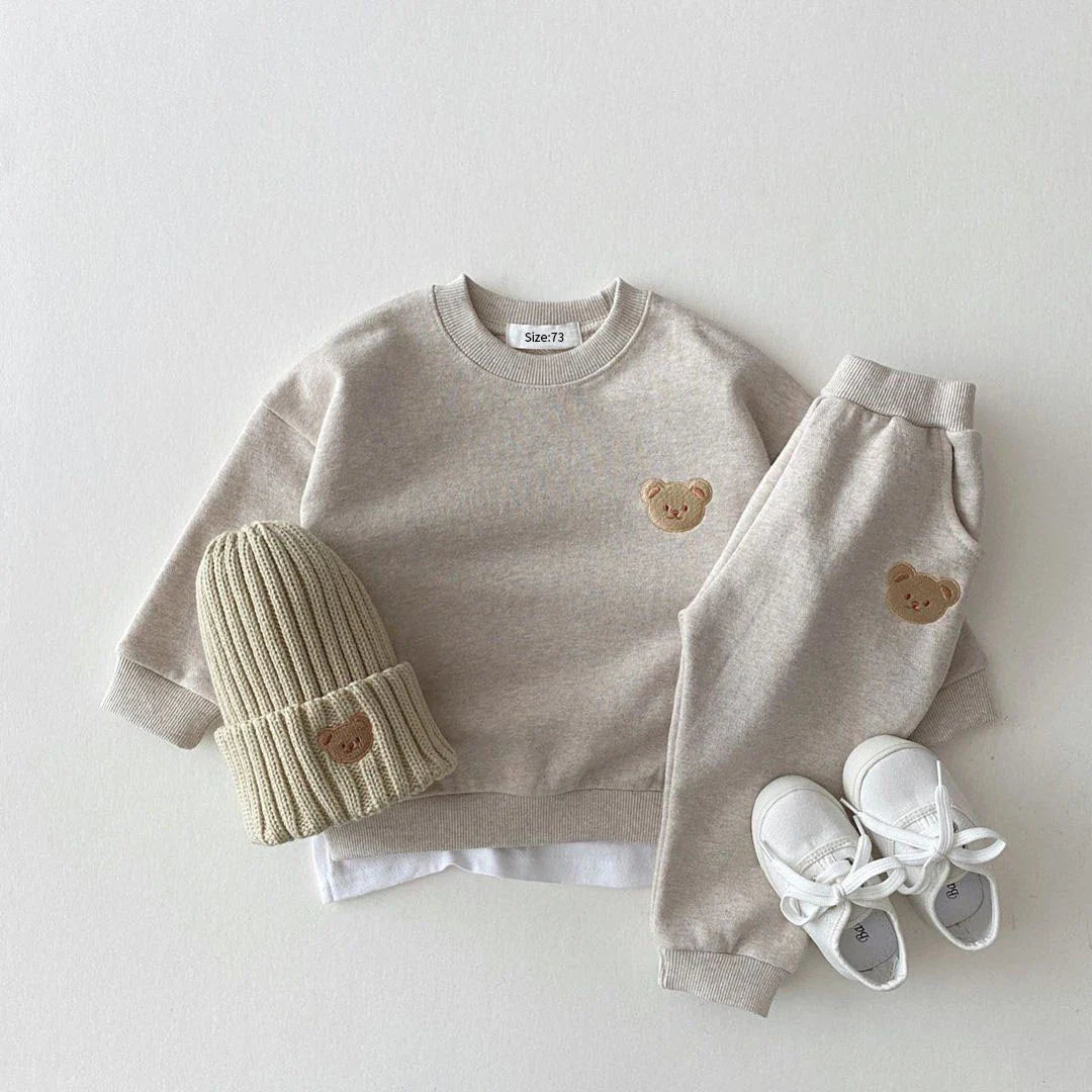 Children's clothing set with gray sweatshirt, pants, white sneakers, and green knit hat on a light gray background.