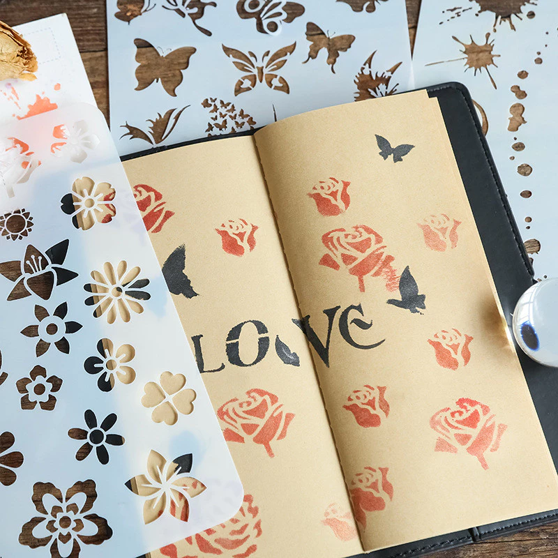 Notebook with floral and butterfly patterns, surrounded by stencils on a wooden surface.