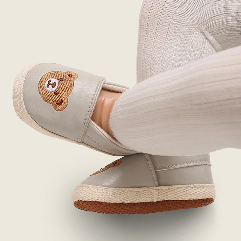 Gray slip-on shoes with a bear emblem worn by a person on a light background