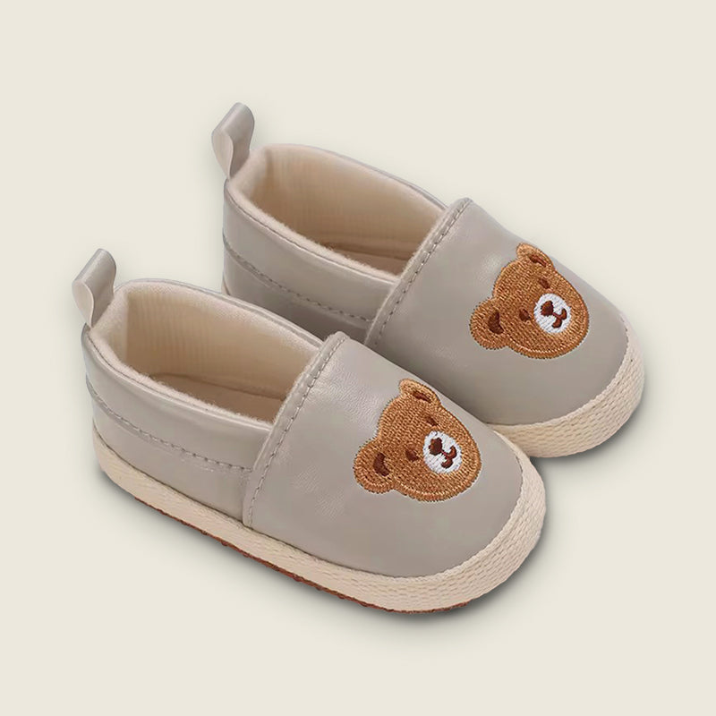 Pair of light gray baby shoes with brown bear faces on a beige background