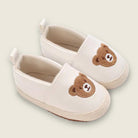 Pair of cream-colored slippers with brown bear faces on a light beige background