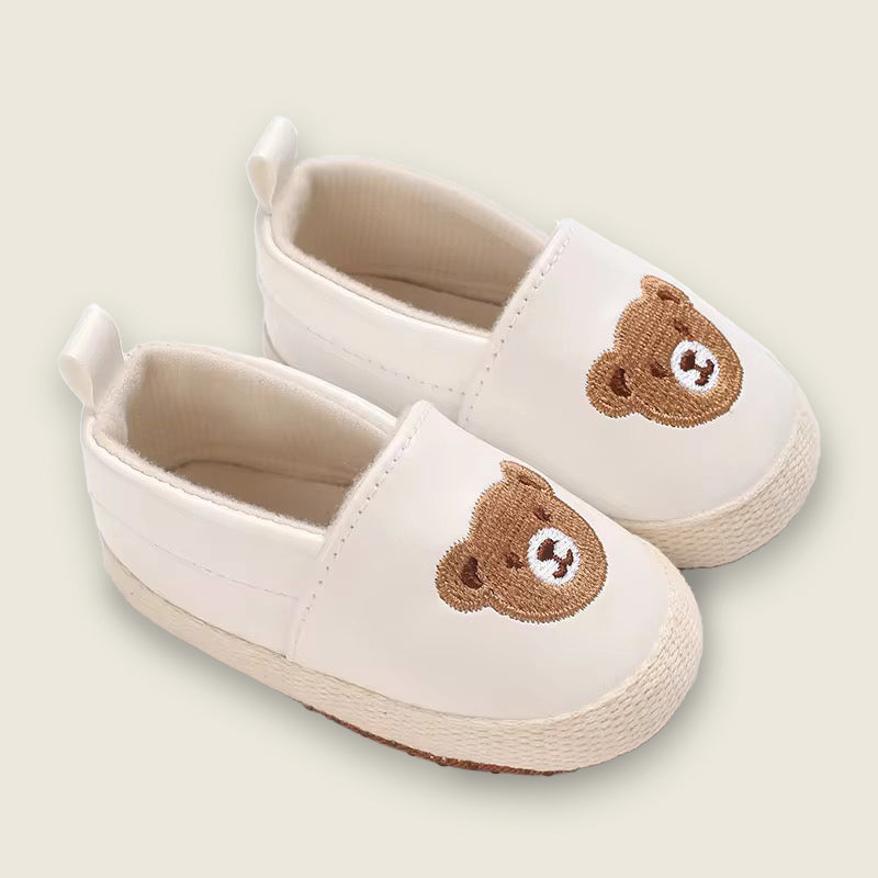 Pair of cream-colored slippers with brown bear faces on a light beige background
